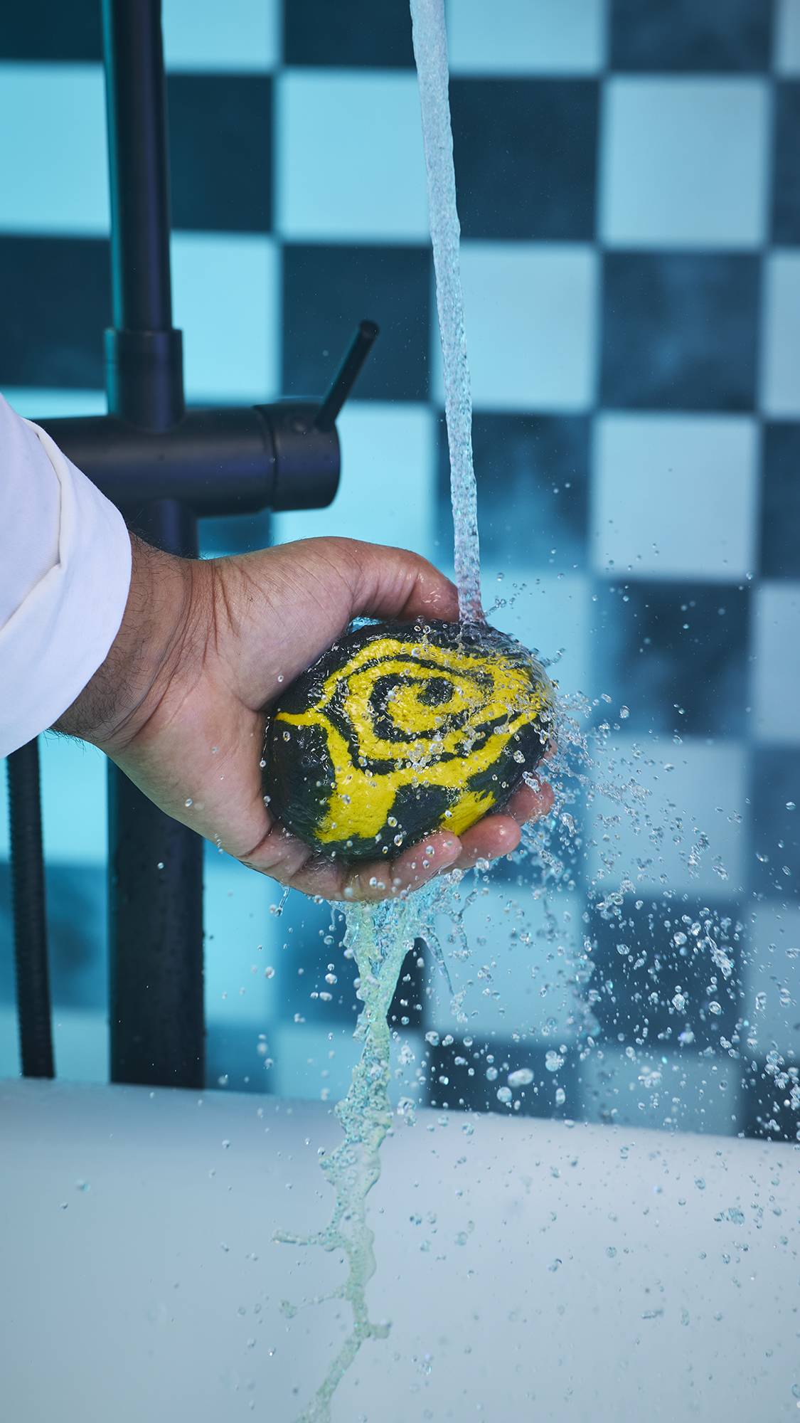 A close-up image of the mode's hand holding the Witchy Woo bubble bar under running tap water. There is a black and white checkered background with a blue hue. 