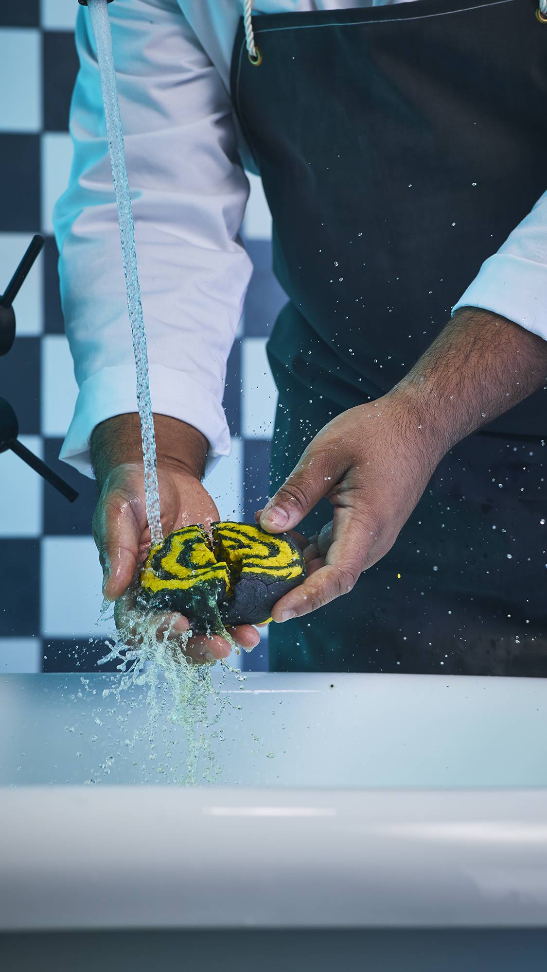 The image shows the model holding the Mini Witchy Woo bubble bar under running water with both hands on a checkered background. 