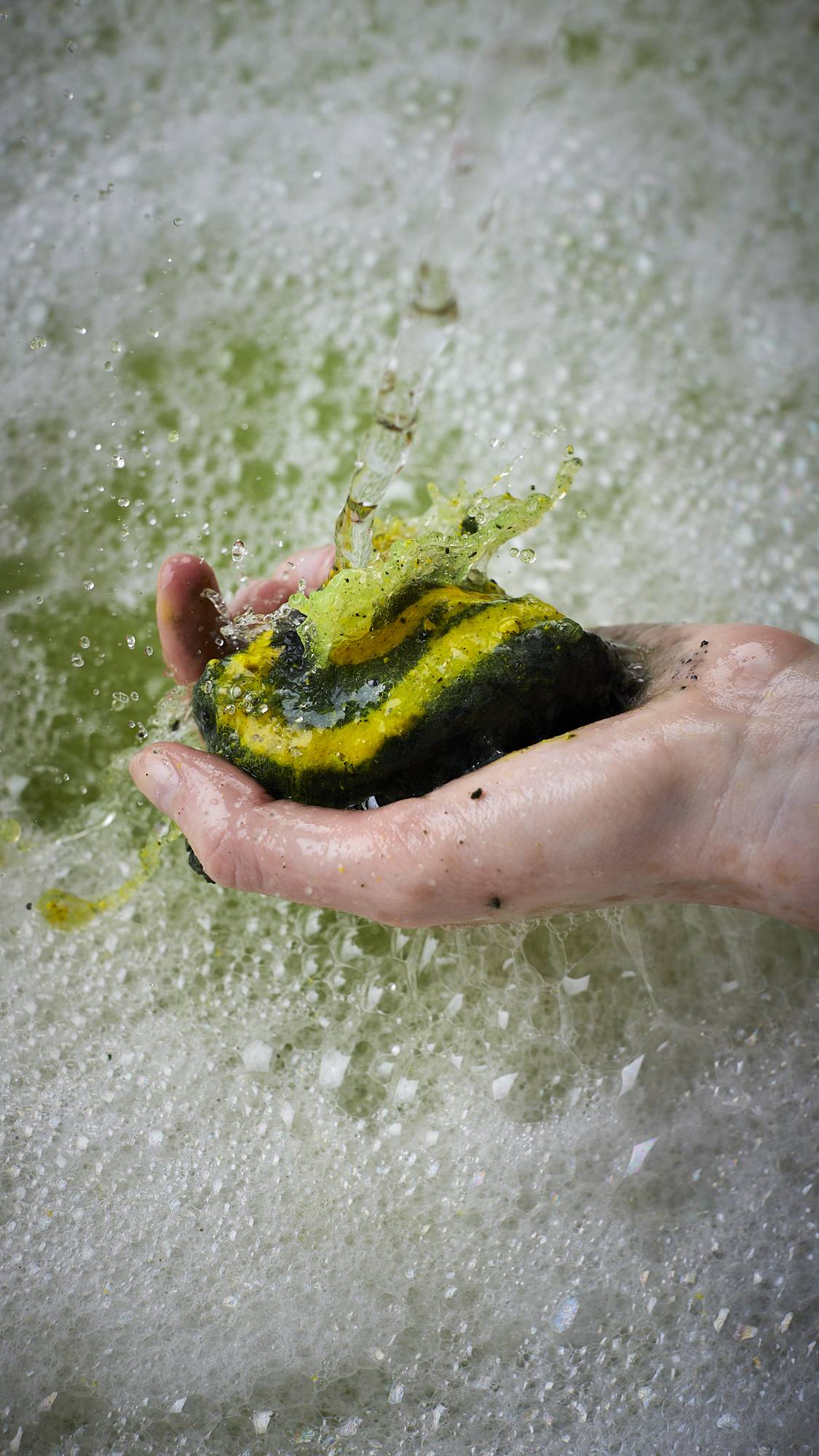 A close-up image of the model's hand holding half of the Witchy Woo bubble bar in their hand under running water. The bath water below is a deep, moss-green colour covered in a layer of bubbles. 