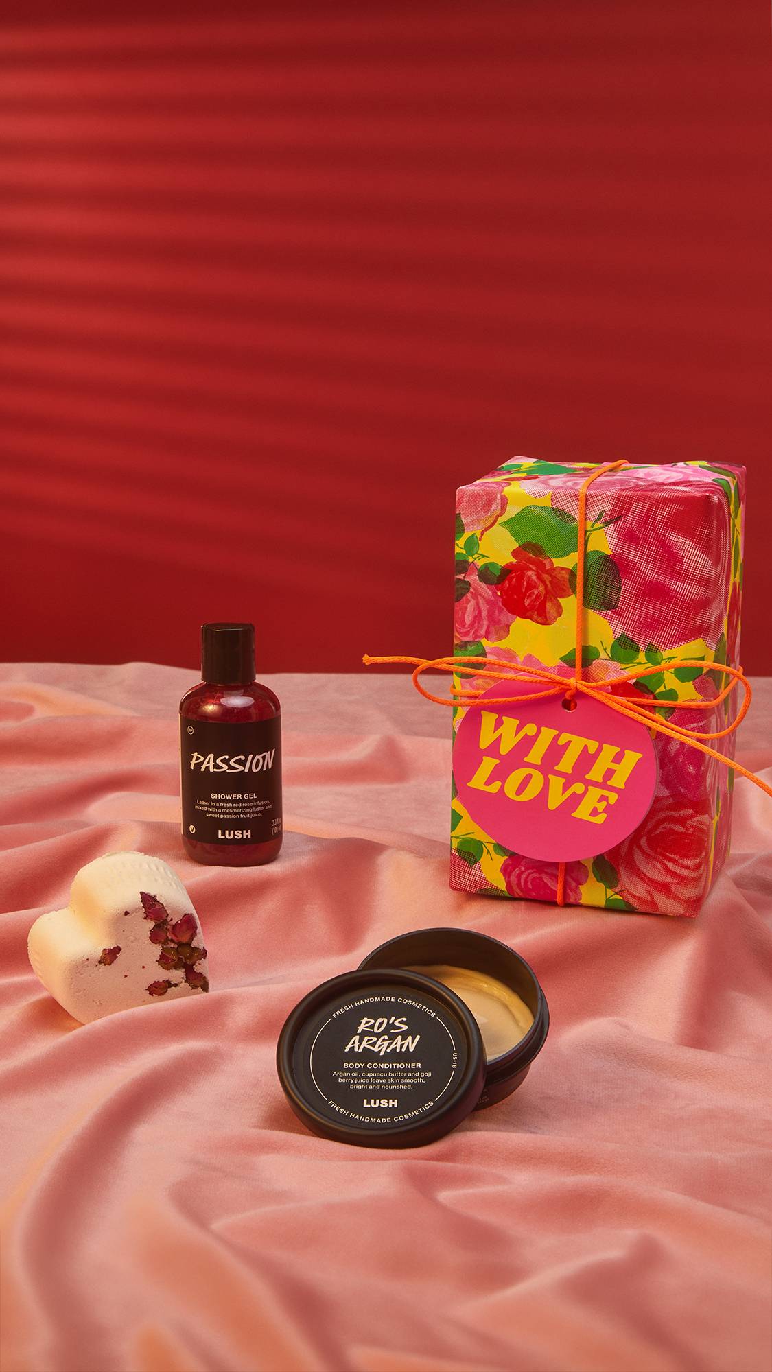 Image shows a red background and a dusky pink satin sheet laid out. The With Love gift box sits on top along with 3 Lush products.