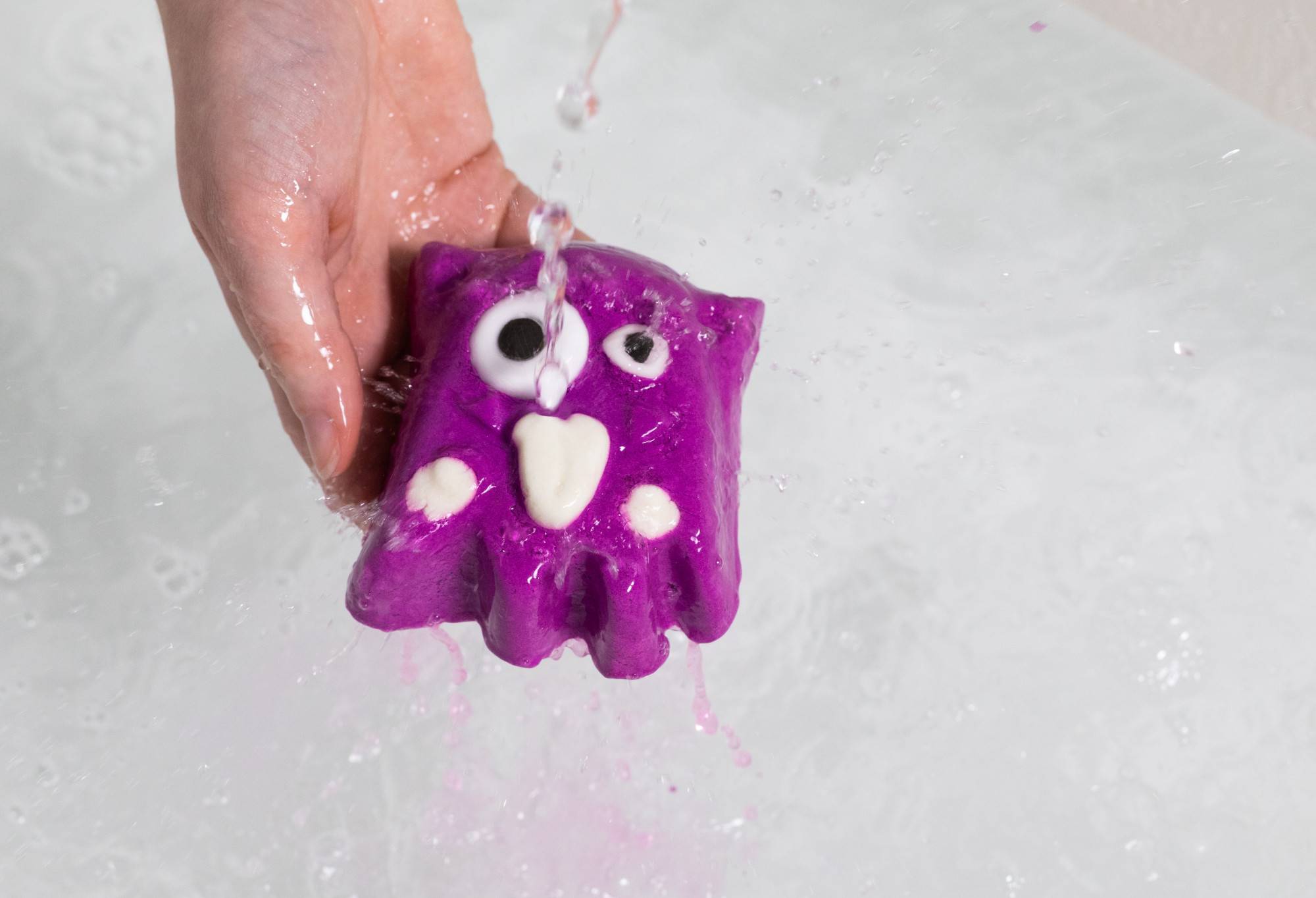 A hand holds the purple Worry Monster under running water. Purple water drops off and into the clear water in the background.