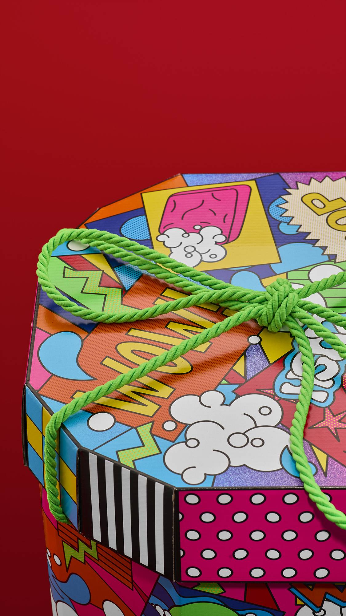 A close-up of a Lush "Wow" gift box, with a vibrant, pop-art-style pattern. The design features colorful comic-book graphics with bold, black outlines, including speech bubbles and the word "WOW," tied with a bright green rope.