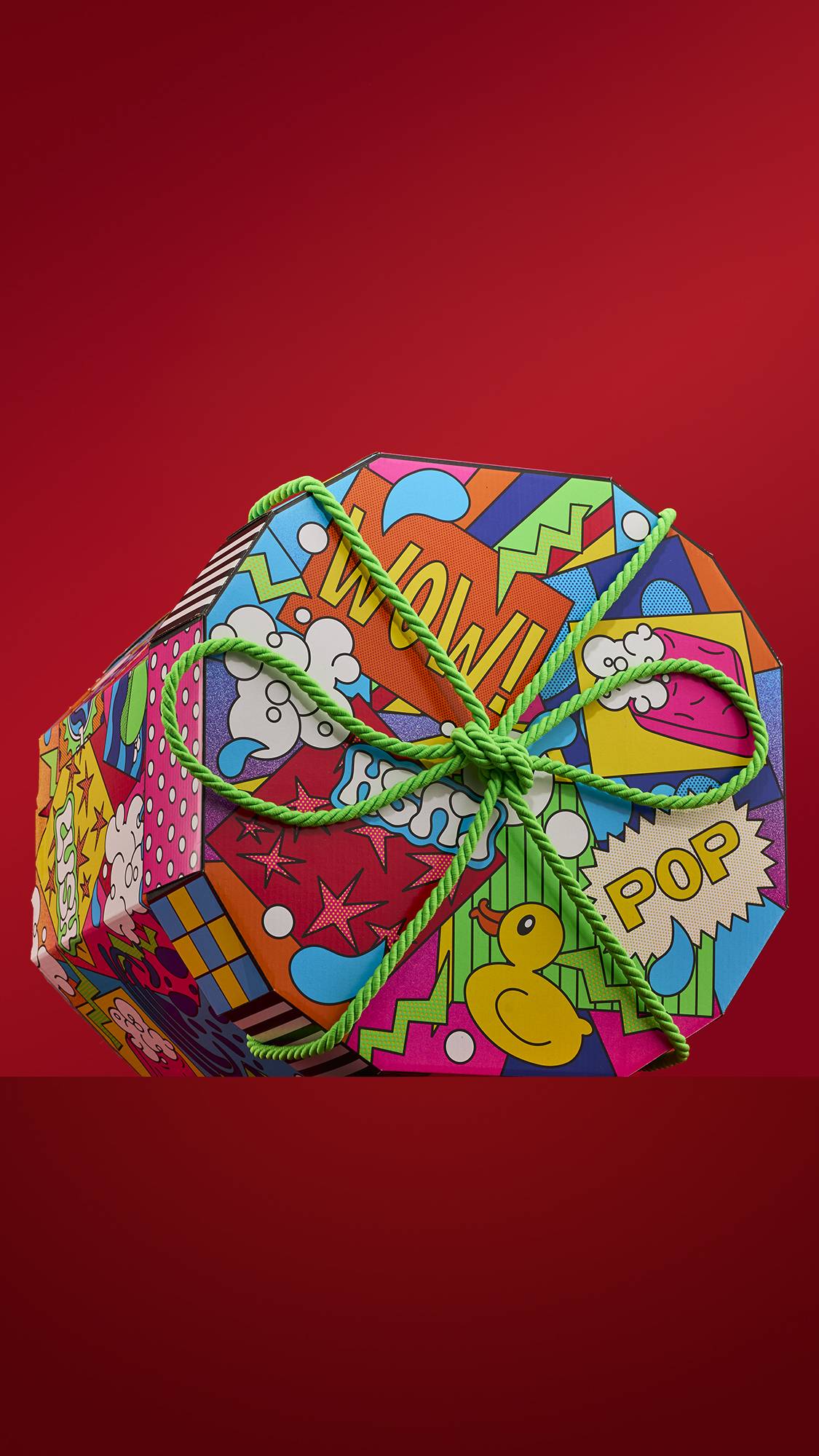 A brightly colored Lush "Wow" gift box with a comic-book-style design. The box is decorated with words like "WOW" and "POP," along with images of rubber ducks and bubbles, and tied with a bright green rope.