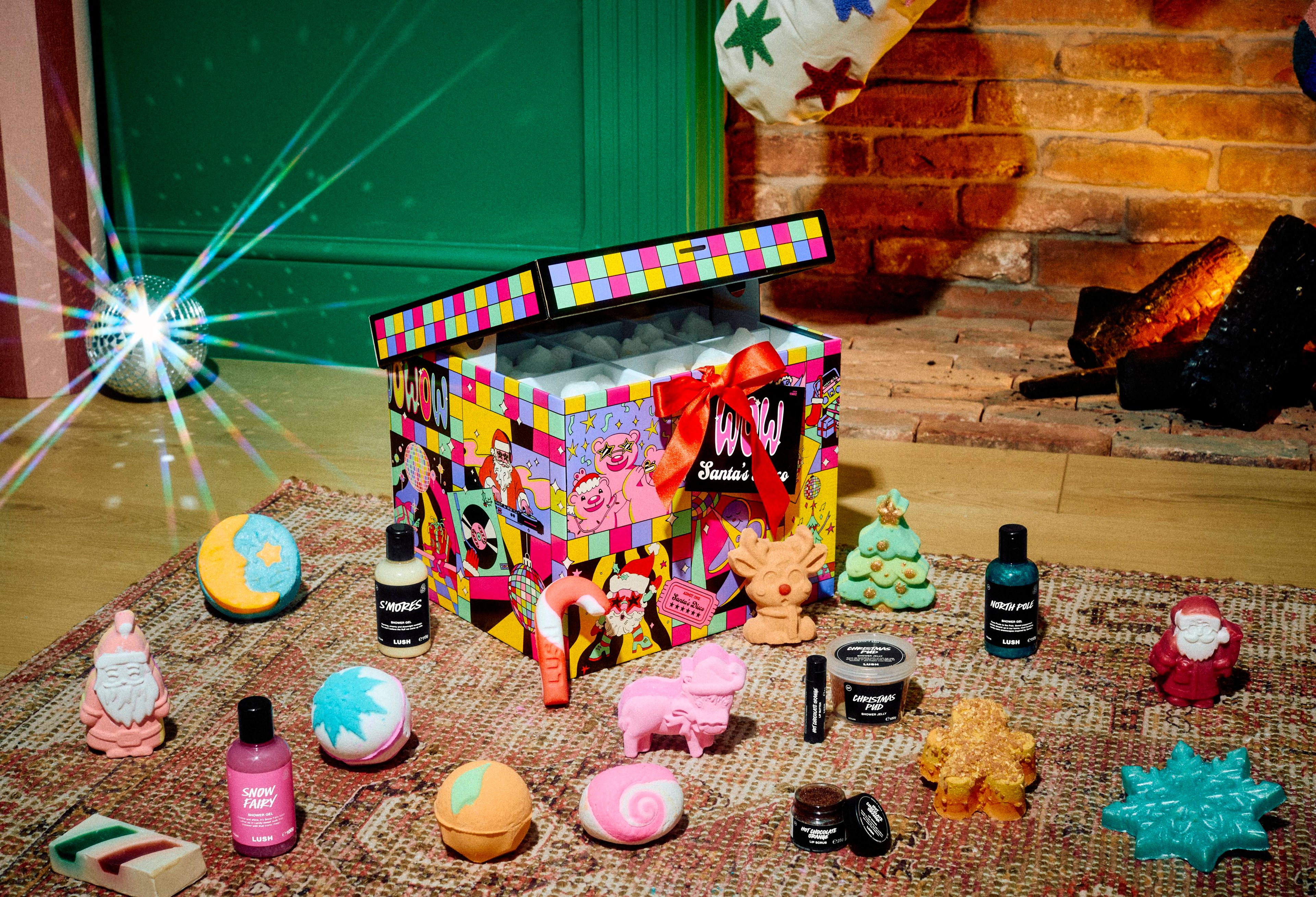 An open Lush "WOW Santa's Disco" gift box sits on a patterned rug surrounded by nineteen assorted festive bath bombs, shower gels, soap, and cosmetics, set in a room with a cosy fireplace and lens flare from a disco ball.