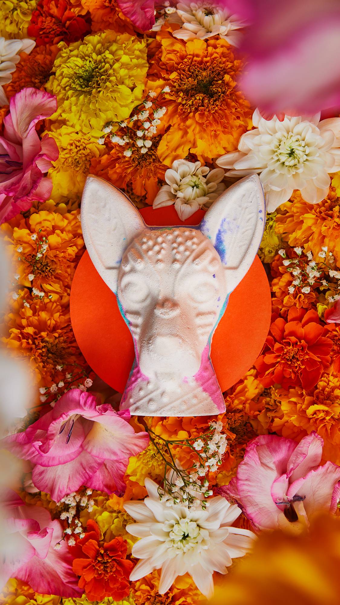 An aerial view image of the Xolo bath bomb sitting on a small, circular orange stand. There are dozens of bright orange, golden yellow and vivid pink flowers surrounding the product. 