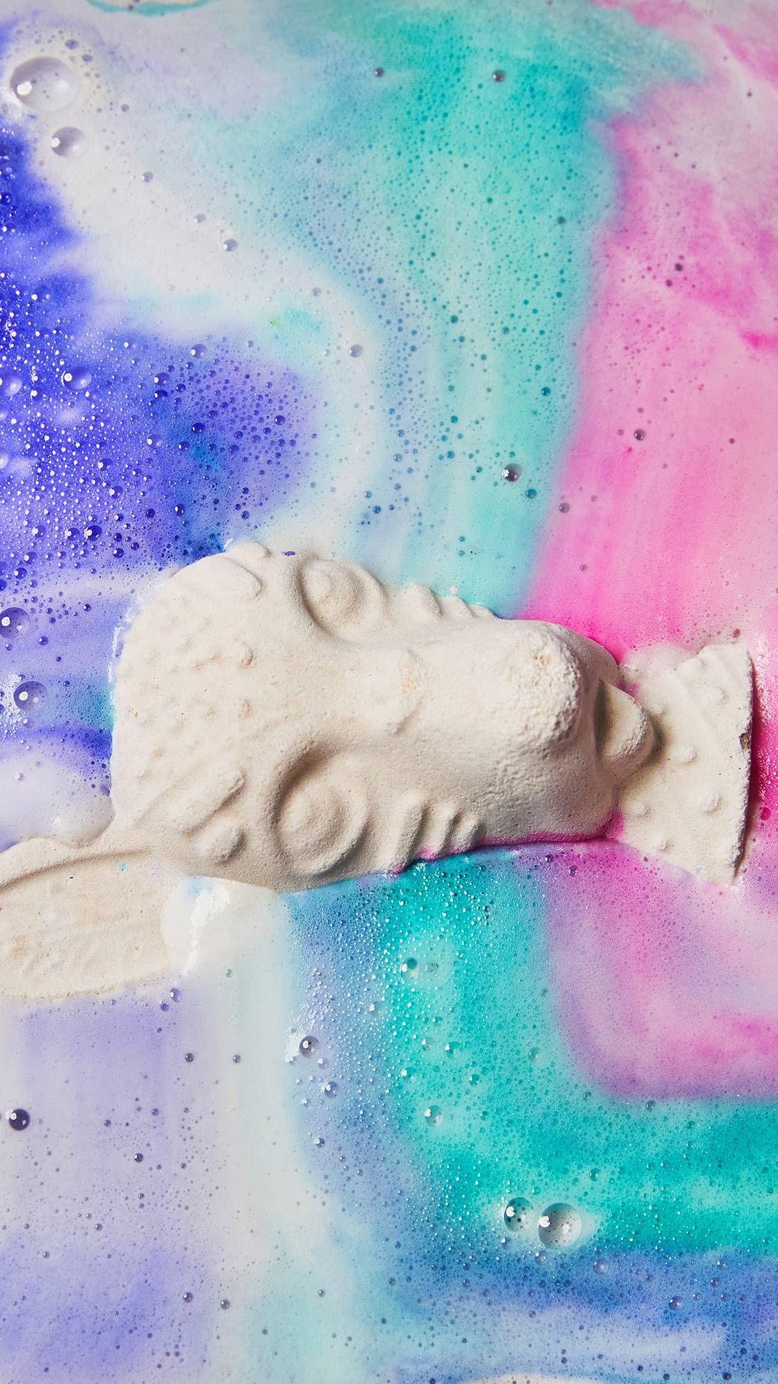 An aerial view of the Xolo bath bomb floating on the bath water as it creates a thick, foamy blanket of blue, purple and pink swirls. 