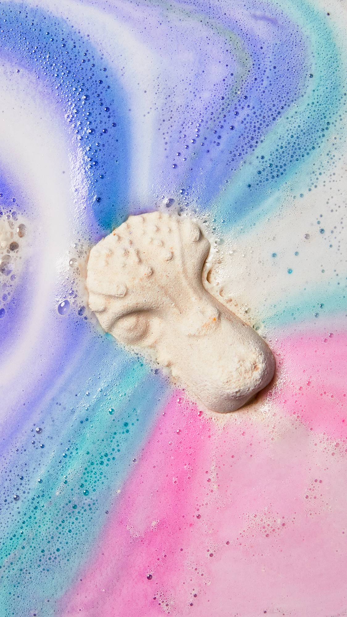 An aerial view of the Xolo bath bomb floating on the bath water as it creates a thick, foamy blanket of blue, purple and pink swirls. 