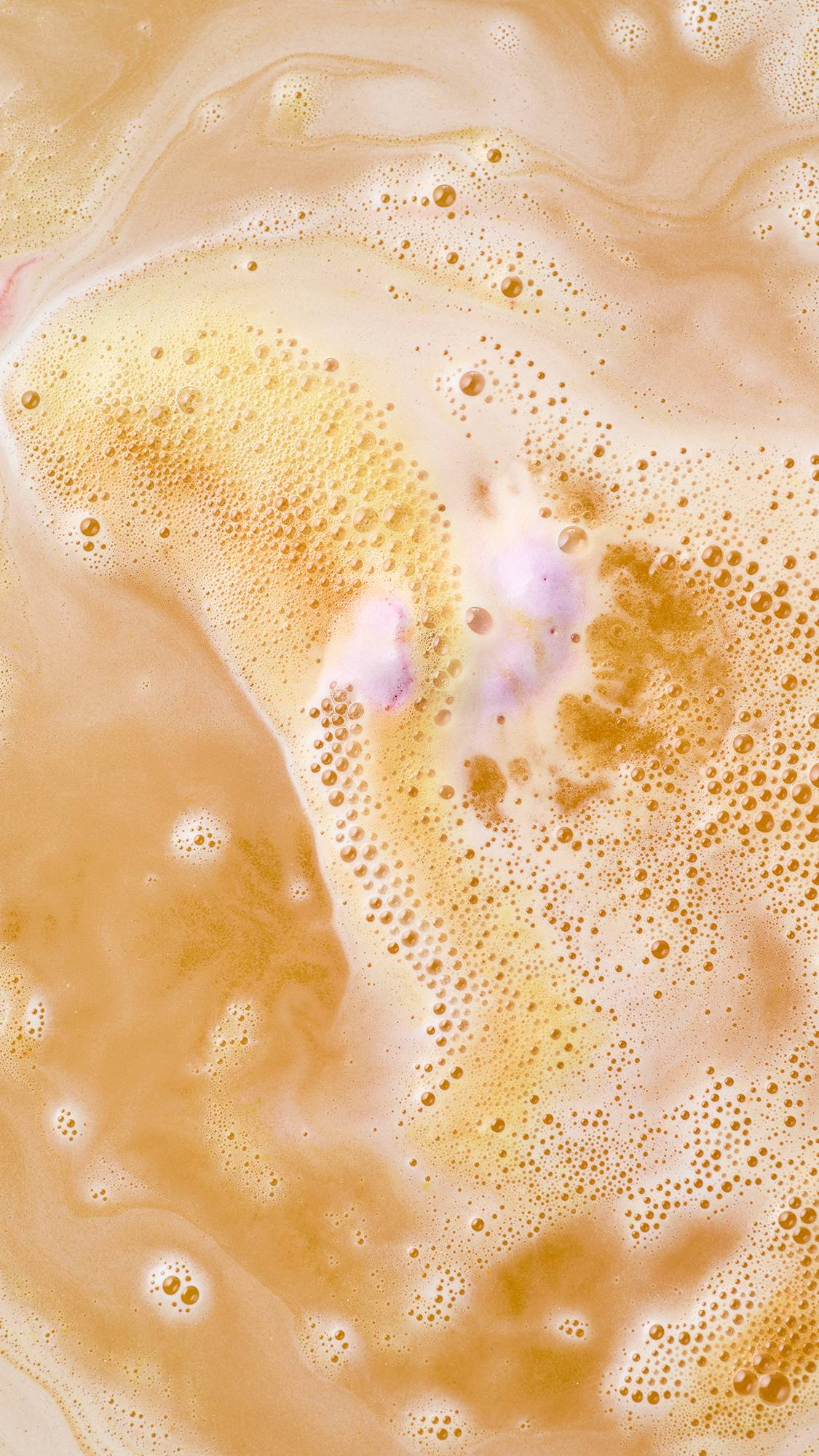 Silky streams of golden-orange and warm peach foam swirl across shimmering bathwater, with delicate hints of pale pink fizz bubbling through the centre as a bath bomb finishes dissolving.