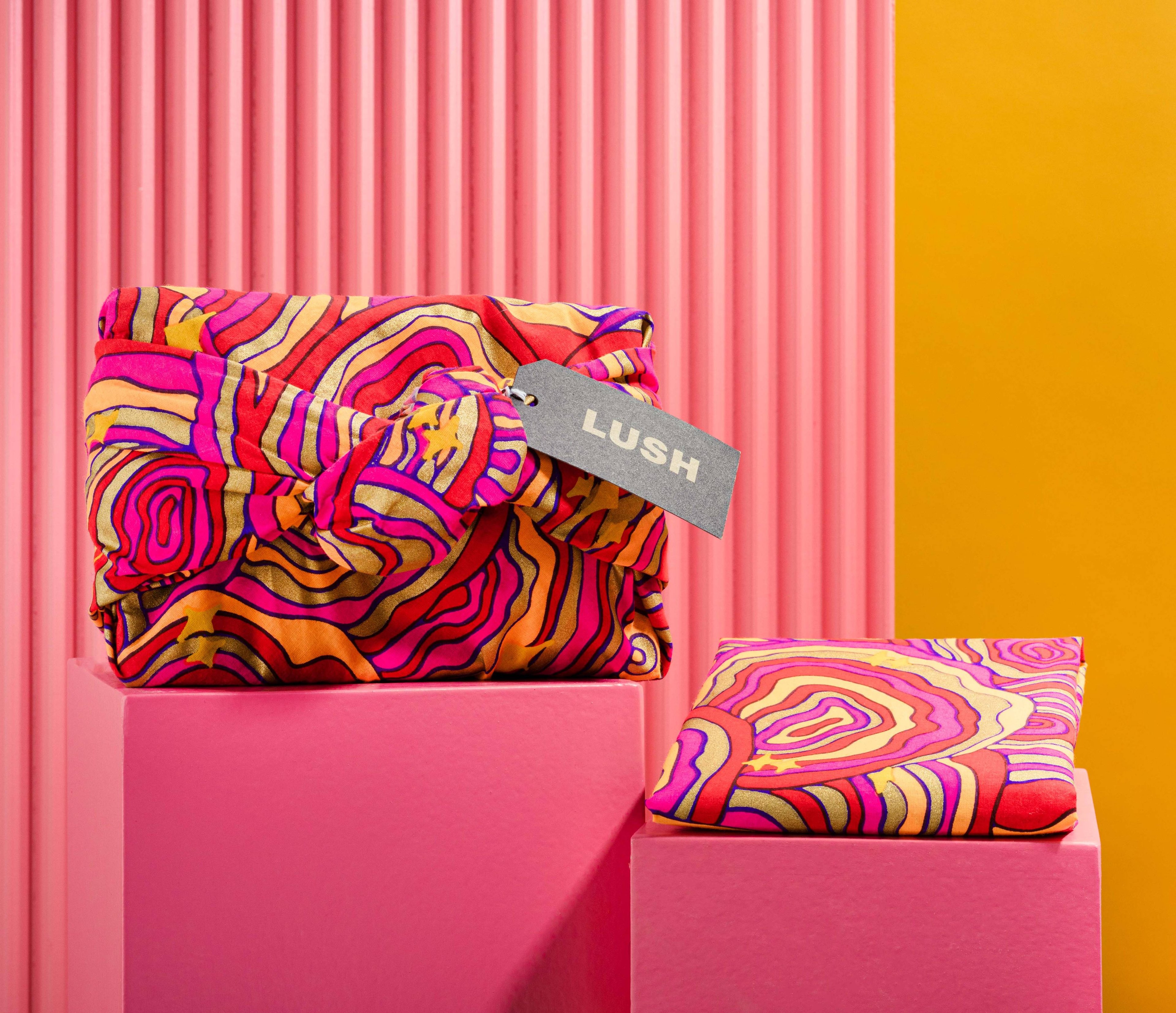 The Knot Wrap is decoratively wrapped with a Lush gift tag, alongside one folded, in front of a pink and yellow background. 