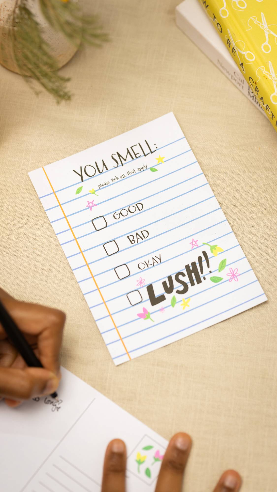 The You Smell: Good, Bad, Okay or LUSH postcard laying on a table as the model writes another card.