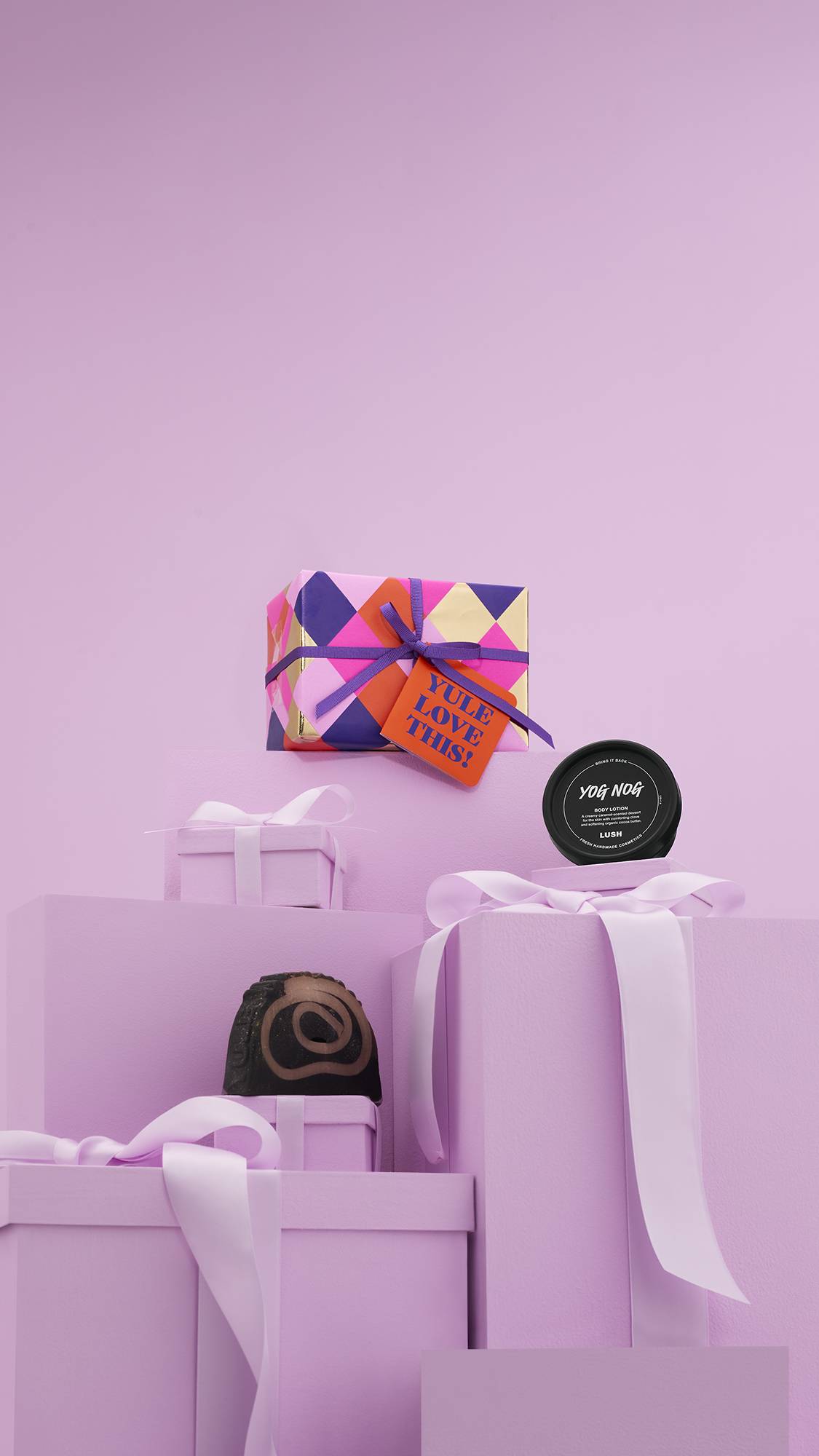 The image shows a large pile of differently-sized wrapped pink gifts with ribbons, on a matching pink background. The vibrant "Yue Love This!" gift box sits on top of the pile, and the two included Lush products sit on other lower pink gifts.