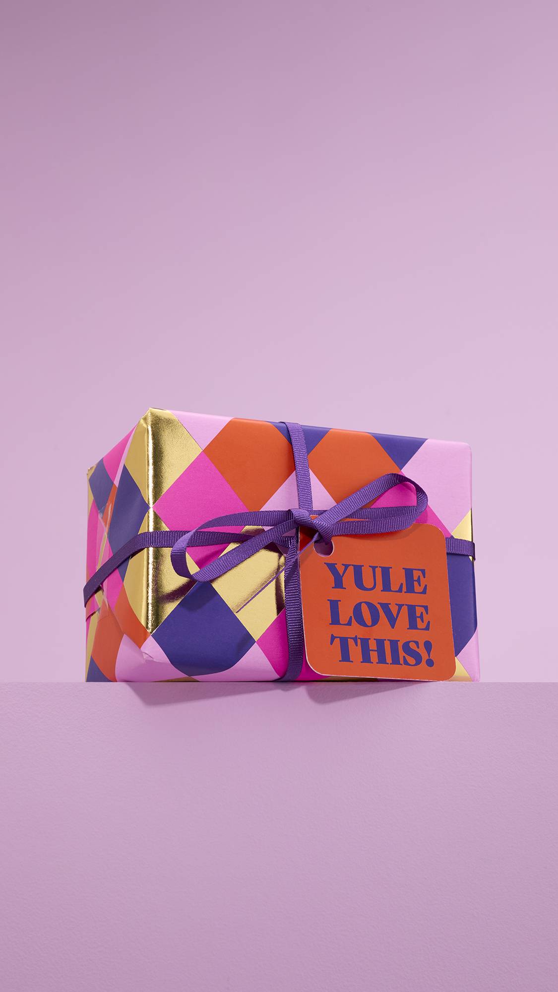 The image shows the vibrant, Yule Love This! gift box, tied with a purple string and an orange gift tag, sitting on a dusky pink platform against a matching pink background. 