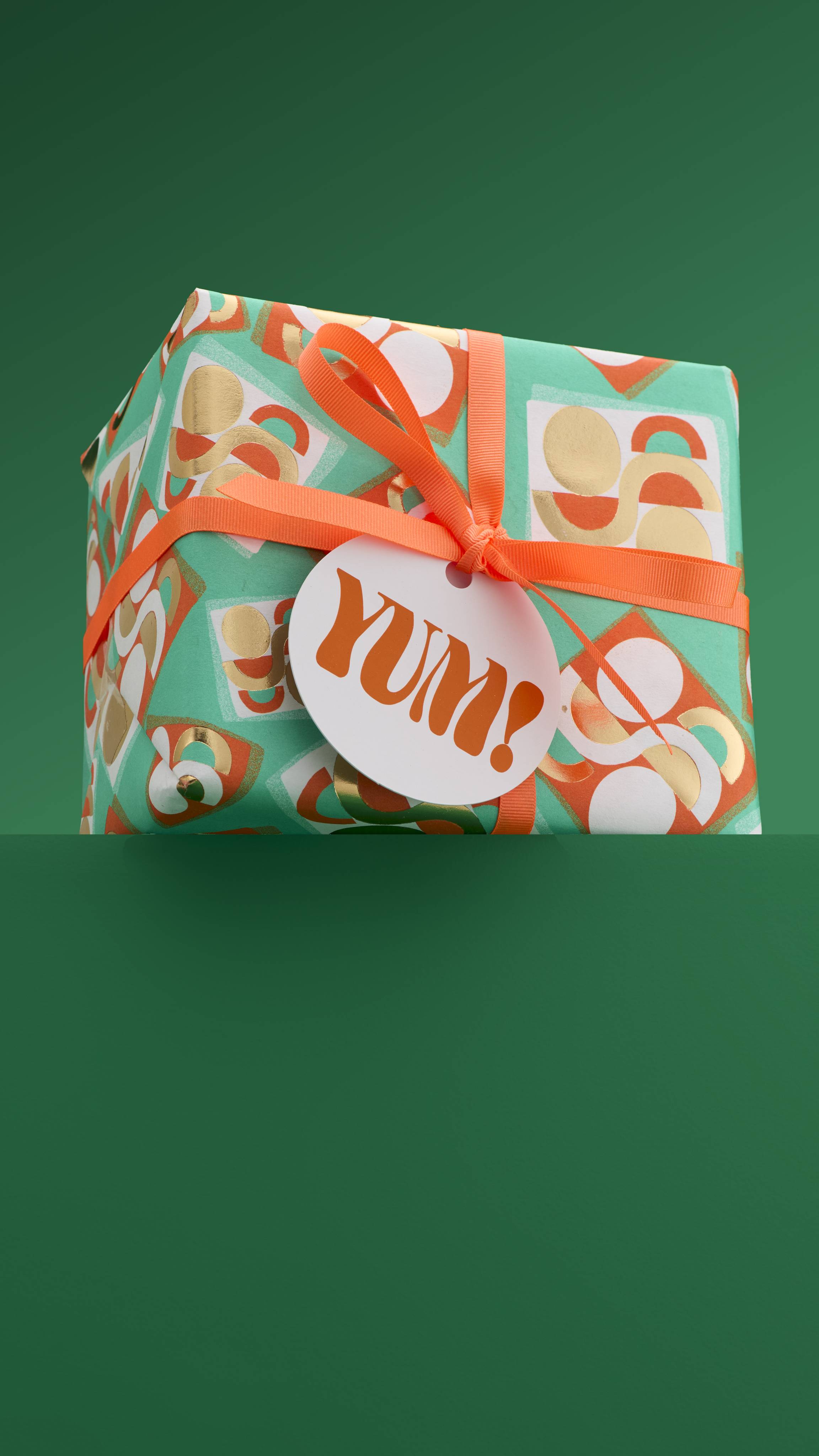 The image shows the colourful, square "Yum!" gift box, tied with orange ribbon and a white gift tag, sitting on a green platform against a matching green background. 