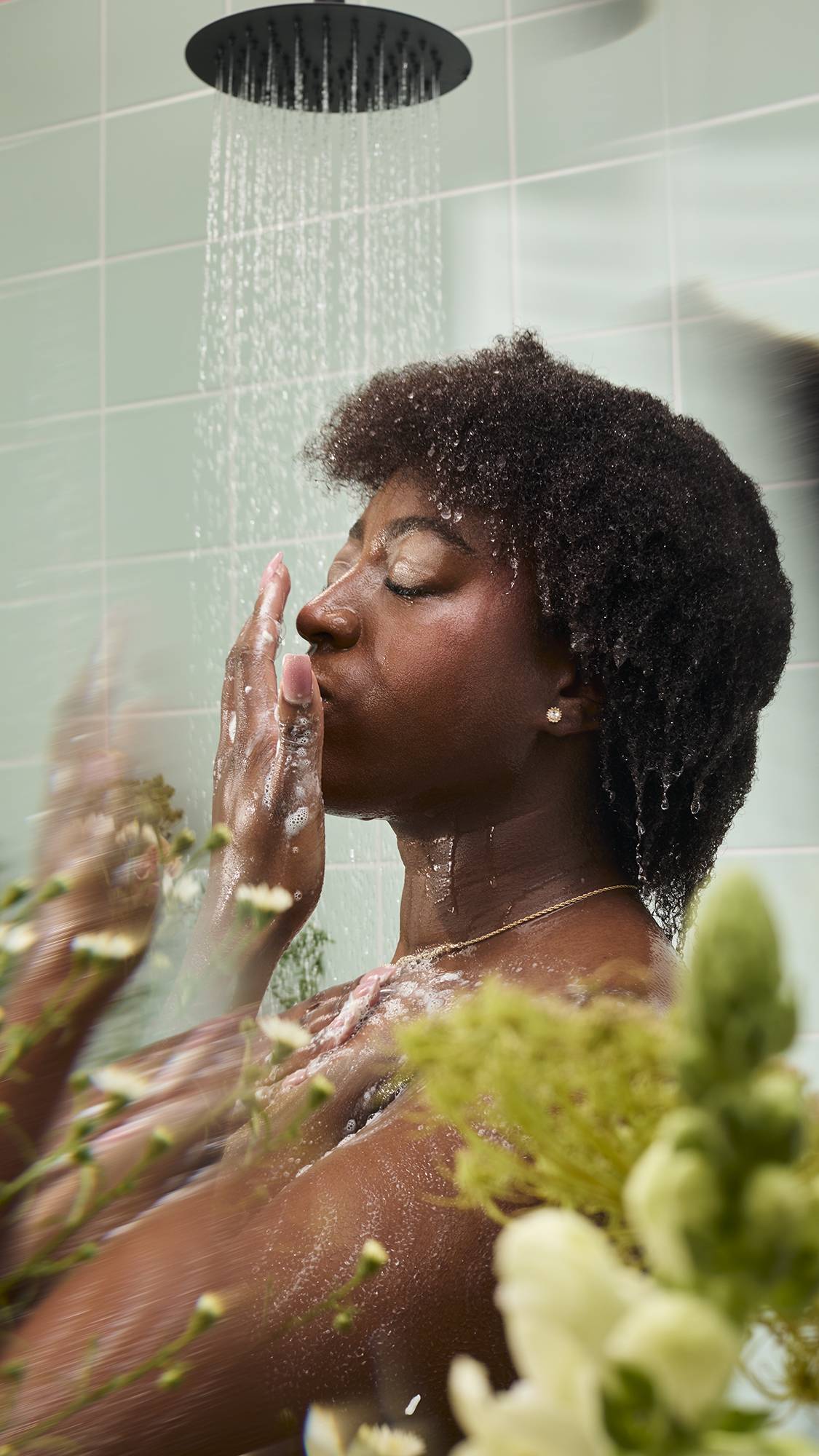 A woman stands under a shower stream, gently washing her face and neck with the "Yummy Custard" body wash. The refreshing scene is framed by blurred flowers in the foreground, set against a backdrop of mint green tiles.