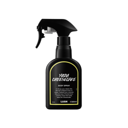 A recycled black plastic LUSH body spray bottle with a trigger nozzle stands upright with a yellow-rimmed black label that reads "Youzu Cheesecake Body Spray". The product description is printed in white text.